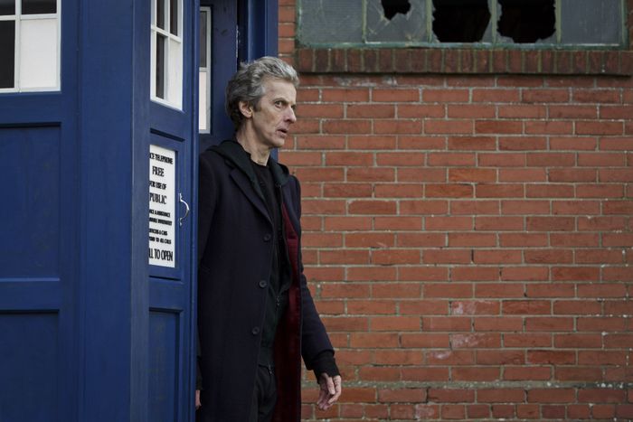 Peter, dressed in a long coat, steps out of the TARDIS, which resembles a police box, against a backdrop of a brick wall.