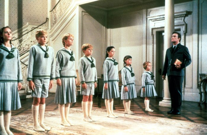 A group of children in sailor-style outfits stands in a line, looking towards an adult holding a book, set in a formal room with a staircase in the background