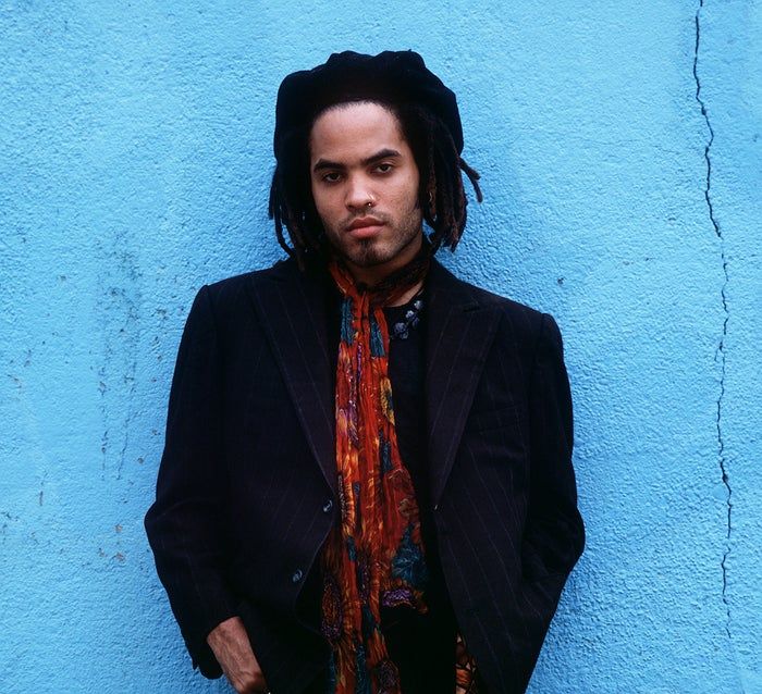A person with dreadlocks, dressed in a pinstripe jacket and a patterned scarf, standing in front of a textured wall