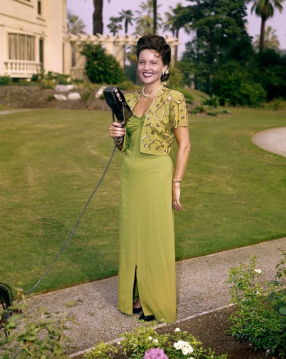 A woman stands outdoors in an elegant vintage gown with detailed top stitching, holding a microphone and smiling, with a garden and building in the background.