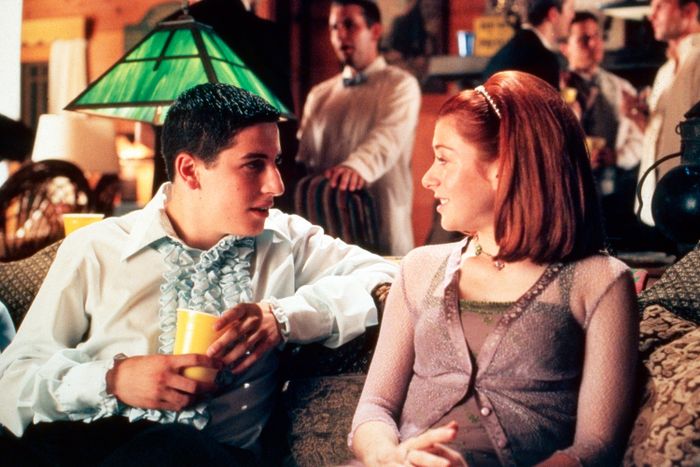 Two individuals lounge on a couch at a party, chatting away. One sports a ruffled shirt, while the other rocks a trendy headband. Party guests mingle in the background