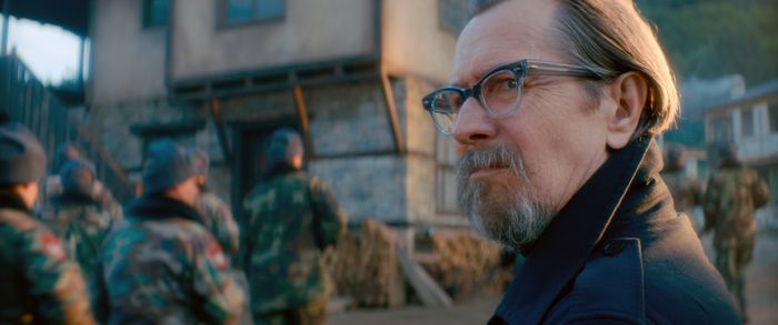 Gary, wearing glasses and a dark coat, stands outside, gazing into the distance as people in camouflage uniforms move around him.