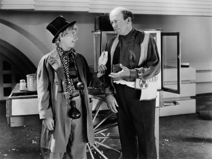 Two individuals in retro clothing stand beside a cart, one wearing a large bowler hat with a clownish appearance, the other in a vest, both engaged in a playful conversation