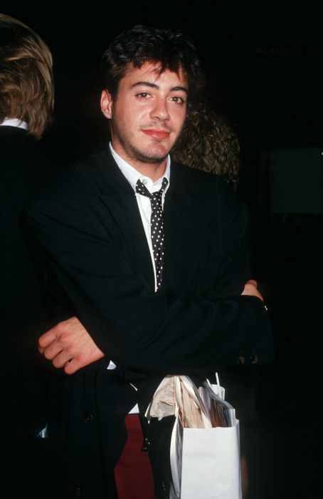 A person dressed in a suit with a polka dot tie, standing with arms folded, holding a paper bag