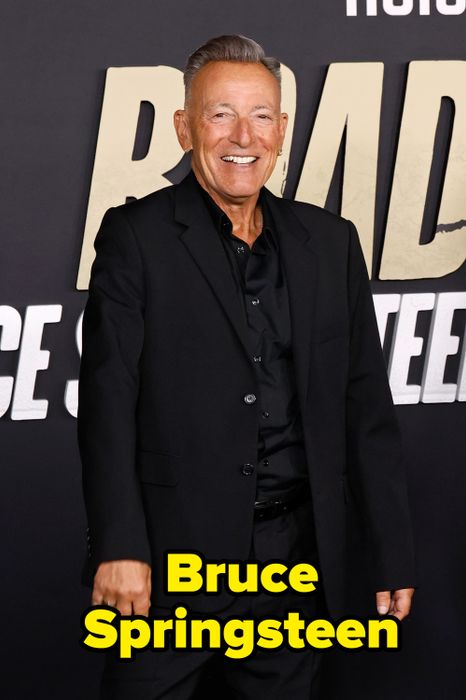 A man dressed in a black suit flashes a smile at a premiere, with bold text in the backdrop