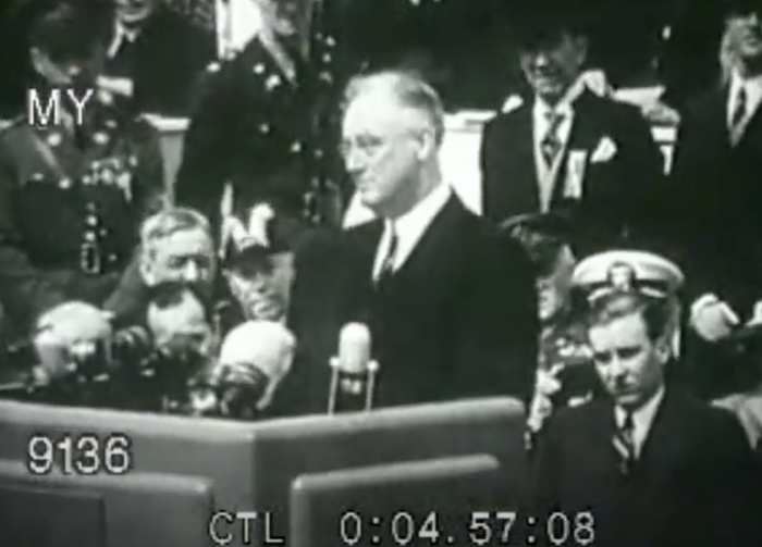 Franklin D. Roosevelt giving a speech at a historical event, standing at a podium with several military and government officials seated behind him.