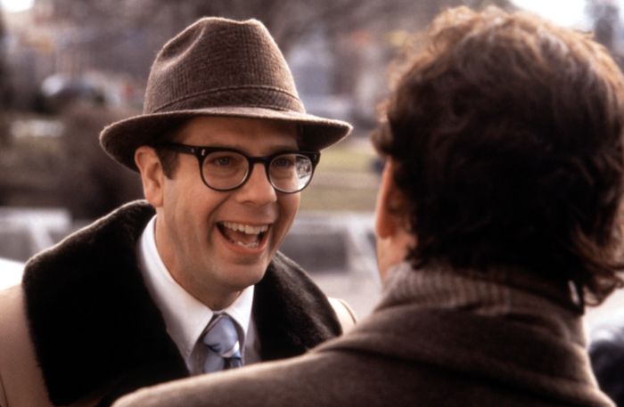 A man wearing glasses and a hat smiles while chatting with another person outdoors.