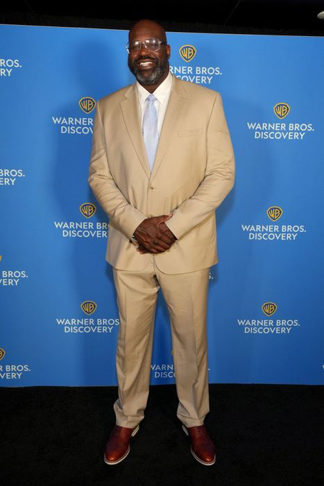 Shaq, dressed in a sharp neutral-toned suit and tie, flashes a smile while posing on the red carpet against a Warner Bros. Discovery backdrop.