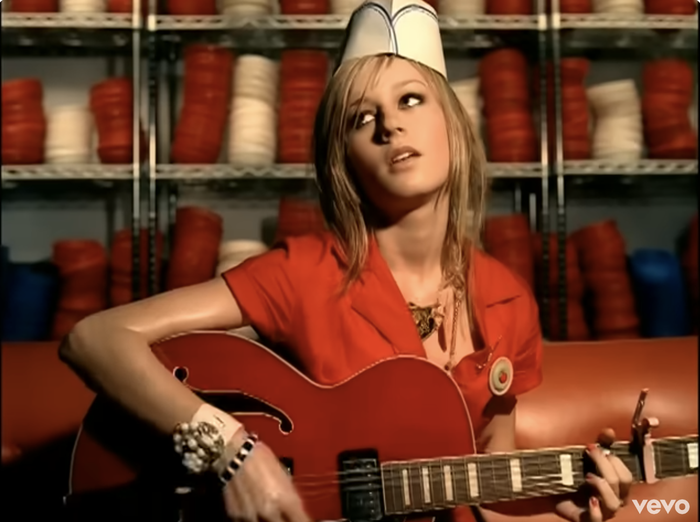 A person dressed in a diner outfit plays a guitar, sitting on a red couch, surrounded by colorful yarn stacks.