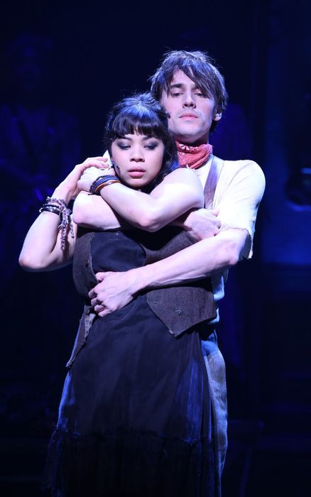 Reeve and Eva share a theatrical moment on stage, with Reeve in rustic attire, wearing a scarf, and Eva in a sleeveless dress, both embodying a dramatic scene.