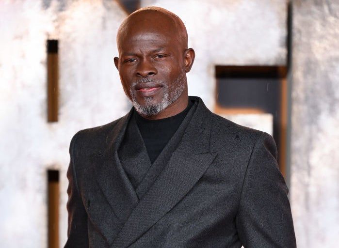 Djimon Hounsou stands confidently on the red carpet in a sharp, dark suit, smiling with his hands in his pockets.