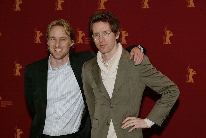 Two individuals on the red carpet. One is dressed in a dark suit and striped shirt, while the other wears a light-colored suit with a scarf.