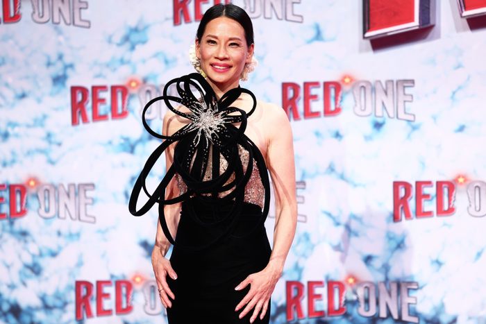 Lucy Liu wearing a sophisticated dress with a detailed, circular fabric design, posing on the red carpet at a media event, with a background featuring 