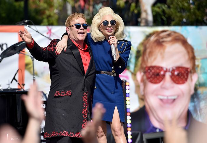 Two performers on stage: one in a decorated suit with sunglasses, the other in a chic dress holding a microphone, both smiling and hugging