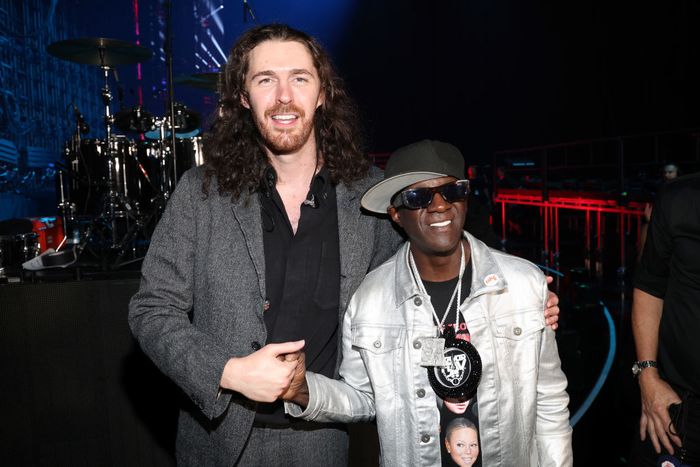 Hozier, in a sleek suit, poses with Flavor Flav, who sports a shiny jacket, hat, and sunglasses at an event. Flavor Flav reaches up to Hozier's shoulder.
