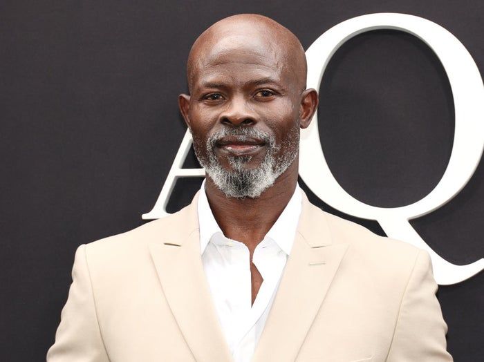 Djimon Hounsou looks sharp in a sleek suit, standing confidently in front of a backdrop with large letters at a star-studded event.