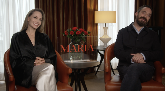 Two individuals sit together, one dressed in a black robe-style top and smiling, next to a man in a jacket, with a table holding roses and the name 'Maria' positioned between them.
