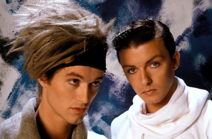 Two individuals with striking hairstyles and fashionable outfits pose for what appears to be a promotional photo or portrait.