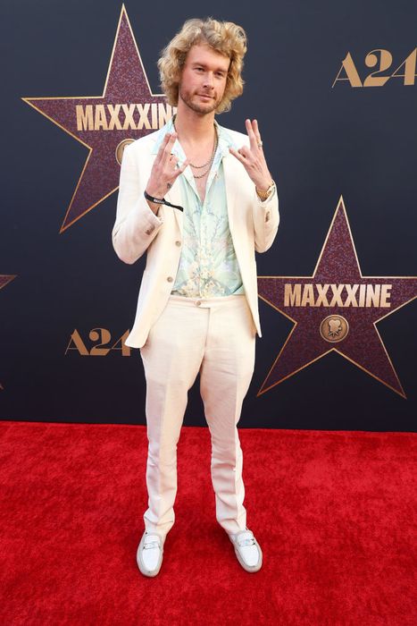 Yung Gravy strikes a playful pose on the red carpet, sporting a sharp suit and a floral shirt, with a starry backdrop behind him.