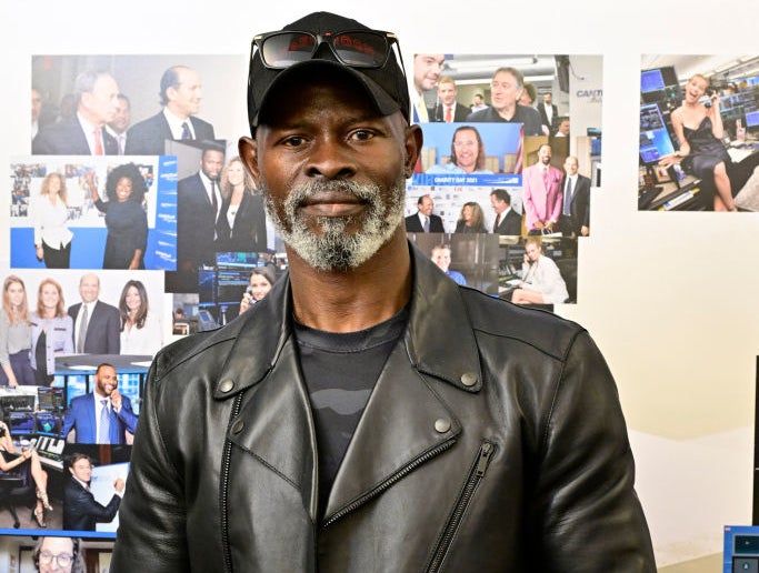 Djimon Hounsou spotted in a leather jacket and jeans at a celebrity event, with various photos and logos appearing in the background.
