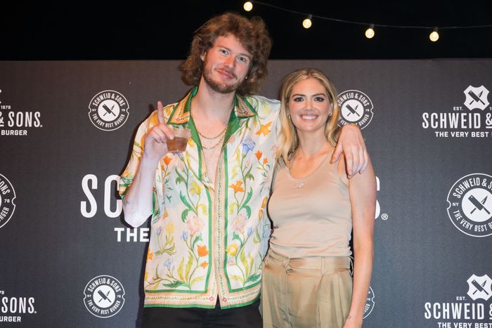 Gravy and Kate pose side by side; he sports a floral shirt, while she opts for a neutral-toned ensemble. They stand before a Schweid & Sons backdrop, with Kate reaching up to his jawline.