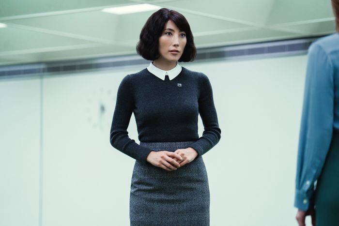 Ms. Casey, in a modern setting, wears a collared sweater with a pin and a patterned skirt, looking engaged in conversation.