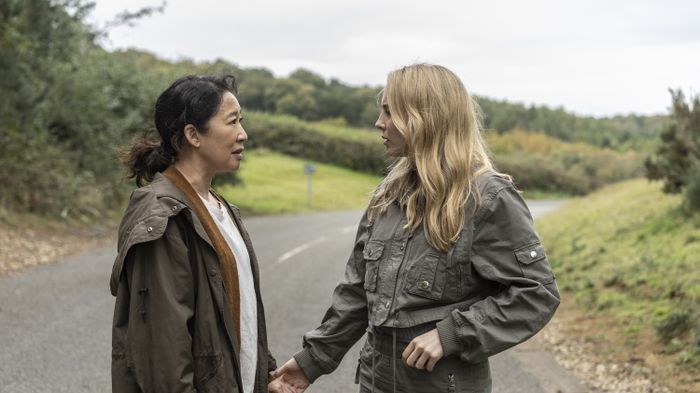 Two individuals stand on a rural road, engaged in a conversation. Both are dressed casually in jackets and seem to be deep in a serious discussion.