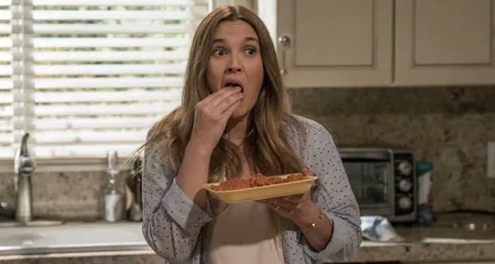 A person sits in a kitchen, eating spaghetti from a plate, looking taken aback. They're dressed casually in a polka-dot shirt with long, flowing hair.