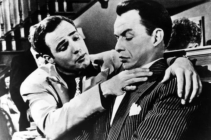 Frank and Marlon in a timeless movie scene; one gently grasps the other's neck. Both wear formal suits, exuding a charged, dramatic atmosphere