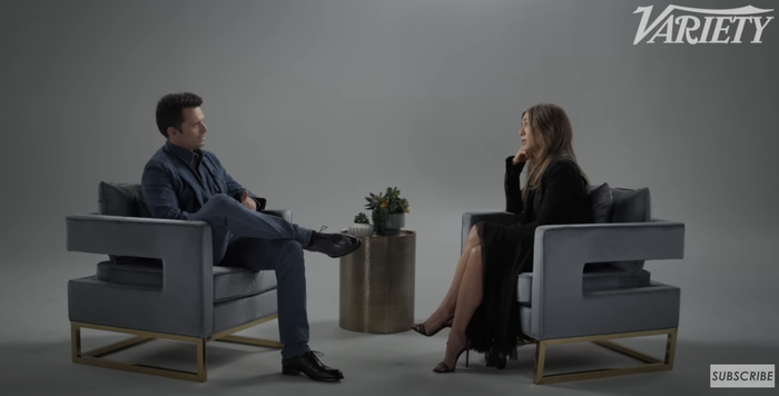 Two people sit in armchairs in a studio, having an interview with a small table adorned with plants between them.