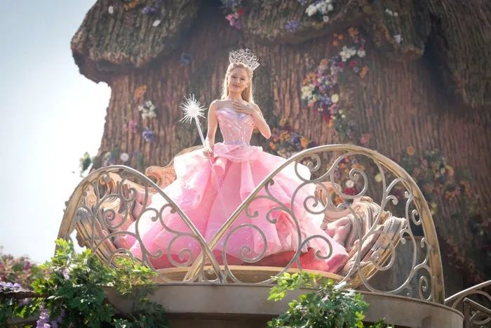 Ariana Grande as Glinda, wearing a fairy tale-inspired gown and tiara, holding a wand while standing on an ornate balcony.