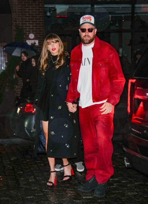 Taylor and Travis walk hand in hand down the street. She sports a chic, embroidered coat, while he wears a striking jacket, pants, and a cap. With Taylor in heels, she reaches just up to his ear.