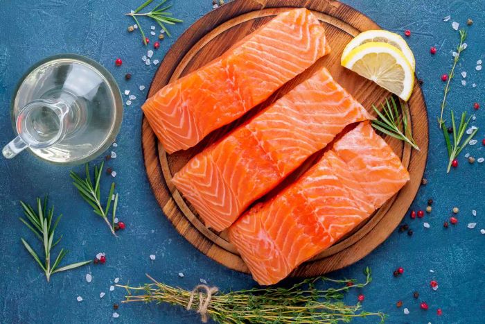A trio of salmon fillets, which are considered a complete protein, placed on a round wooden cutting board with fresh lemon slices alongside.