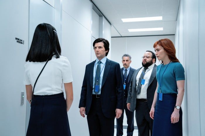 A group of five professionals in formal attire stand in a corridor, focusing intently on a woman with long hair facing them.