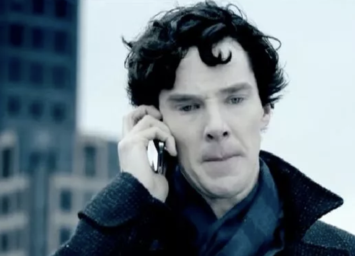 A man with curly hair, dressed in a textured coat, holds a phone to his ear and appears lost in thought against a cityscape backdrop.