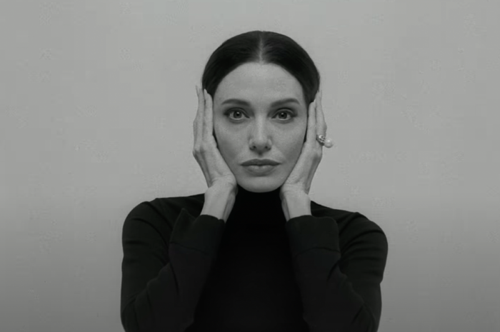 A person wearing a turtleneck poses with hands framing their face, set against a plain background.