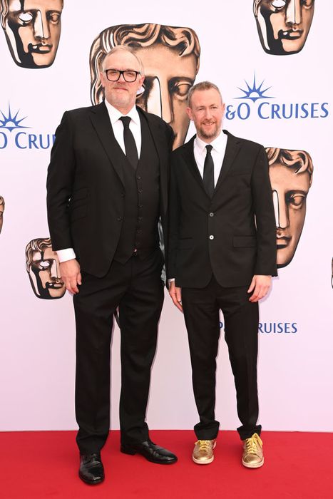 In the spotlight at a red carpet event, Greg and Alex pose in suits, with Alex sporting gold sneakers. The backdrop features a 'Cruises' theme with BAFTA mask designs, and Alex's height reaches just up to Greg's ear.