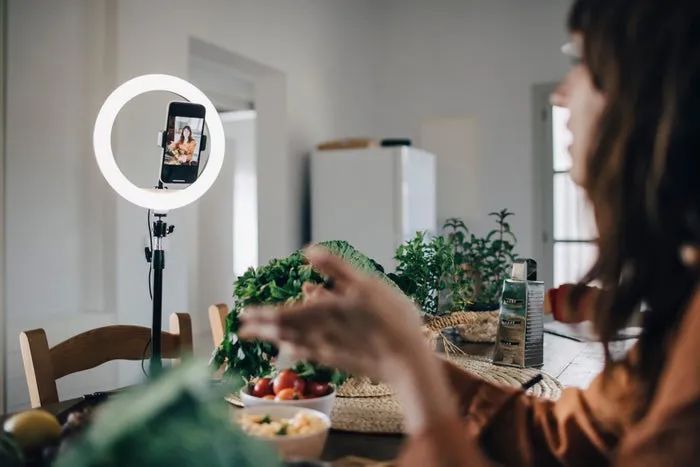A person filming a cooking video at a kitchen table, surrounded by fresh ingredients, with a ring light in use