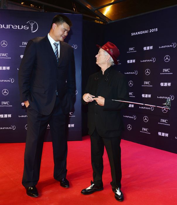On the red carpet, Yao wears a suit while Bill sports a formal outfit with a distinctive bright hat. The two are caught in a conversation, with Bill falling short of reaching Yao's shoulder.