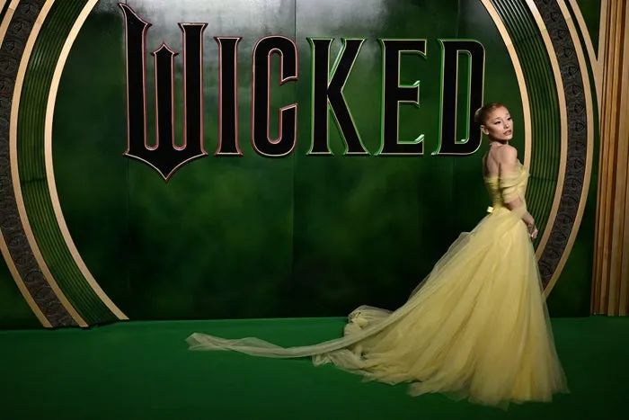 Ariana Grande strikes a pose on a green carpet, dressed in a stunning strapless gown with a flowing train, with the 'Wicked' logo displayed prominently in the background.
