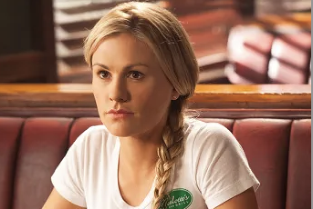 A person sitting in a diner booth, wearing a white t-shirt and sporting a braided hairstyle, gazing forward with a neutral expression.