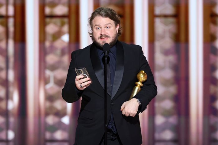 Brady Corbet, dressed in a suit, holding an award statue and a phone, stands in front of a microphone at an event.