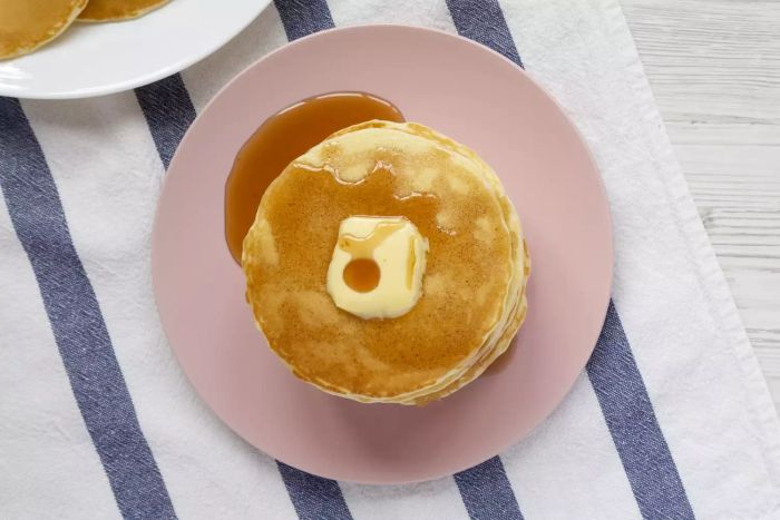 A close-up shot of a stack of homemade pancakes topped with butter and drizzled with maple syrup, served on a pink plate. Viewed from above.