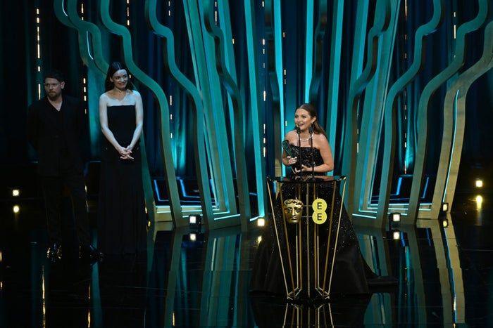A trio of individuals stand on a stage, one delivering a speech at an awards event. The group is dressed in sophisticated formal attire.