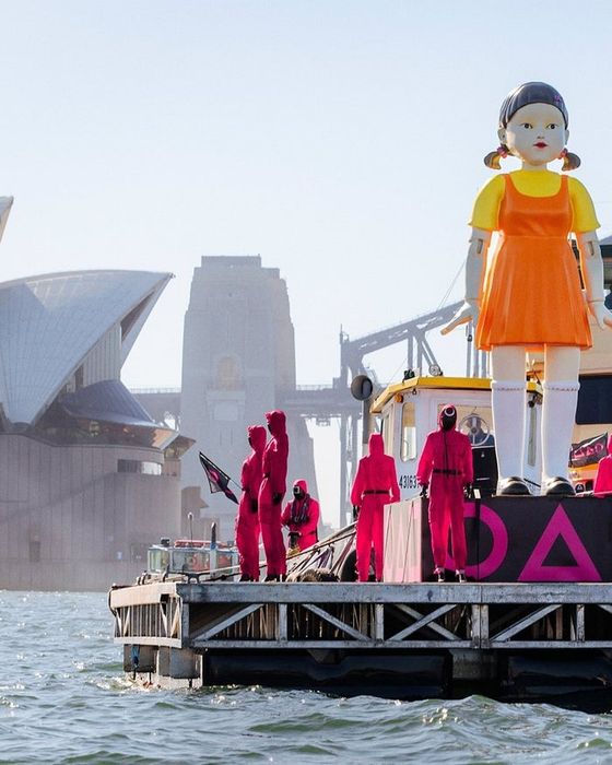 A massive doll and individuals in pink costumes are seen on a barge in front of the iconic Sydney Opera House.