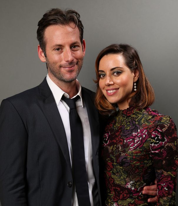 Jeff Baena, dressed in a suit and tie, stands beside Aubrey Plaza, who is wearing a floral high-neck dress. Both are smiling for the camera against a simple backdrop.