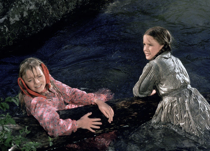 Two young girls in vintage dresses are joyfully holding onto a log, smiling, as they float down a serene creek. They seem carefree and full of happiness.