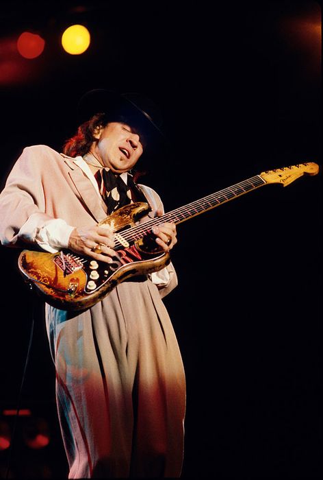 A musician passionately plays an electric guitar on stage, dressed in a loose suit and a wide-brimmed hat.