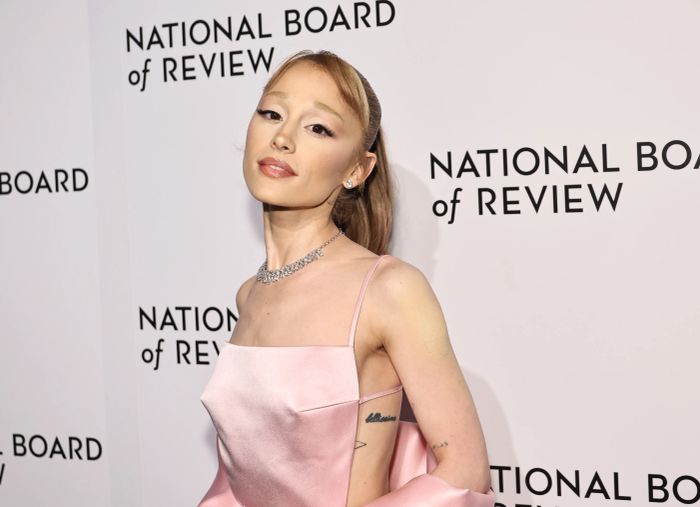 Ariana Grande poses at a National Board of Review event in a satin pink dress, with a visible shoulder tattoo.