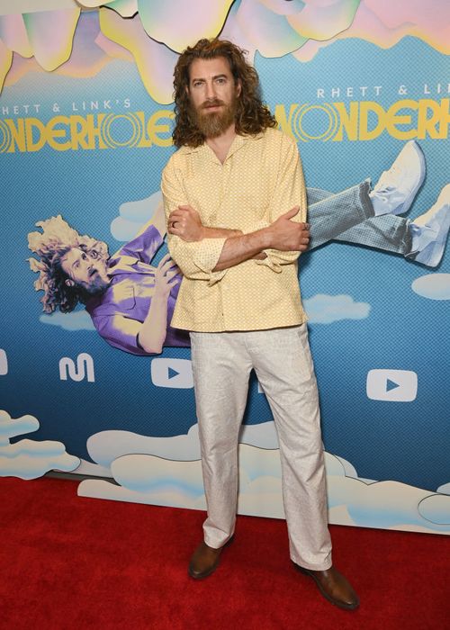 Rhett steps onto the red carpet in a bold patterned shirt and light-colored pants, posing in front of a playful, eye-catching poster backdrop.